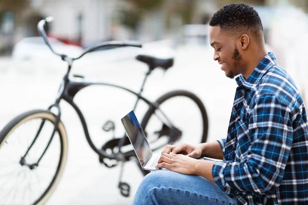 Homem preto usando laptop, sentado na cidade com bicicleta — Fotografia de Stock