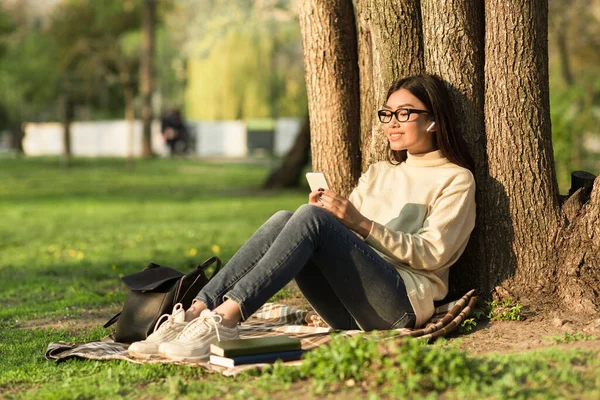 Студентка використовує телефон, сидячи під деревом в парку — стокове фото