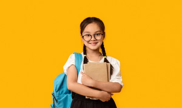 Vrolijk japans schoolmeisje knuffelen boek staande over gele achtergrond — Stockfoto