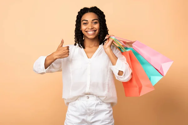 アフリカ系アメリカ人の女の子ジェスチャー親指アップ保持ショッピングバッグ、ベージュの背景 — ストック写真