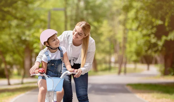 Anne kızına bisiklet sürmeyi öğretiyor. Hafta sonunu parkta geçiriyor.