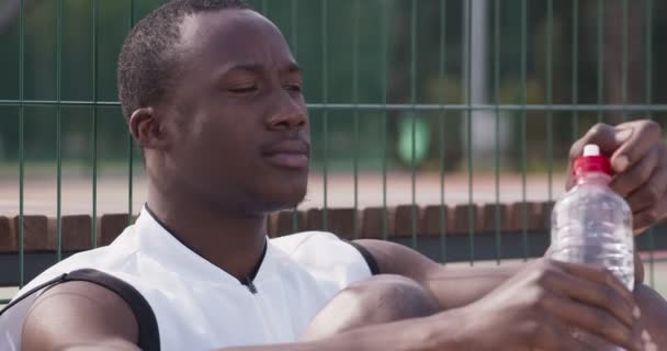 アフリカ系アメリカ人の男が水を飲んでバスケットボールのスポーツグラウンドに座って — ストック動画