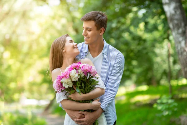 Celebrando el aniversario. Chica joven con flores mirando afectuosamente al novio en el parque — Foto de Stock