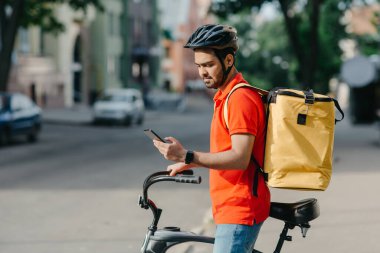 Modern mobil uygulamalar. Kasklı, çantalı, bisikletli genç adam akıllı telefona bakıyor.