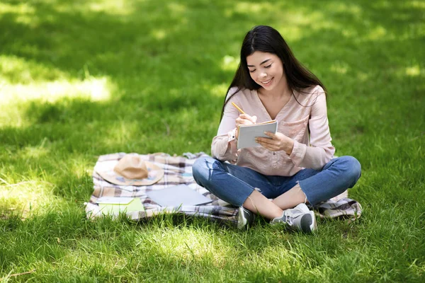 Krásná asijská dívka sedí na kostkované v parku, dělat si poznámky do notepadu — Stock fotografie