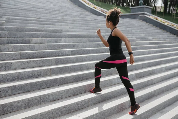 Moda menina americana africana em sportswear com fones de ouvido e rastreador de fitness corre ao longo das escadas da cidade — Fotografia de Stock