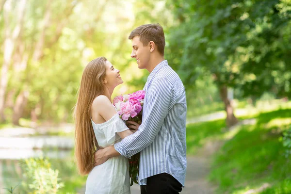 Chica encantadora con ramo de peonías abrazando a su novio en una cita romántica en el parque —  Fotos de Stock