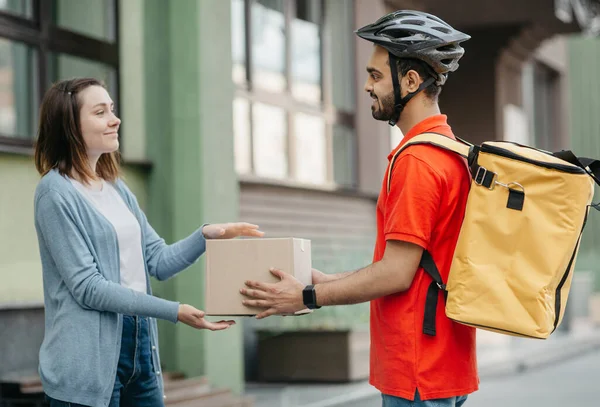 Glückliche Frau erhält Paket. Lieferant in Helm mit gelbem Rucksack gibt Box an Kunden, in der Nähe von Haus — Stockfoto