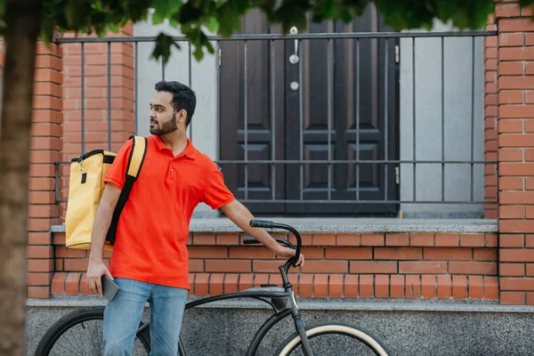 Ev teslimatıyla birlikte modern online alışveriş. Sarı sırt çantası ve bisikleti olan, sakallı, ciddi bir genç adam. — Stok fotoğraf