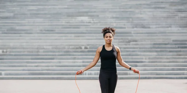 Spor kıyafetli, kablosuz kulaklıklı, spor kulaklıklı, fitness takip cihazı takan merdivenlerde ipe atlayan Amerikalı bir kadın. — Stok fotoğraf