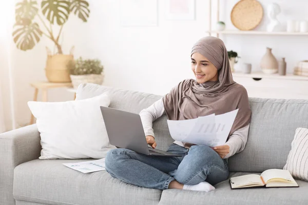 Trabajo desde el concepto del hogar. Mujer musulmana sonriente ocupada con computadora portátil y papeles — Foto de Stock