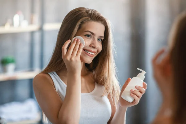 Adorável jovem mulher usando removedor de maquiagem ou loção facial na frente do espelho no banheiro — Fotografia de Stock
