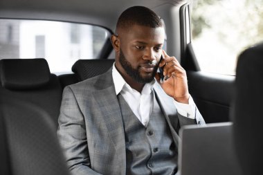 Genç Afrikalı iş adamı arabada cep telefonu ve laptop kullanıyor.