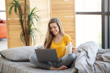 Neşeli genç kız evde çalışmak ya da online çalışmak için dizüstü bilgisayar kullanıyor.