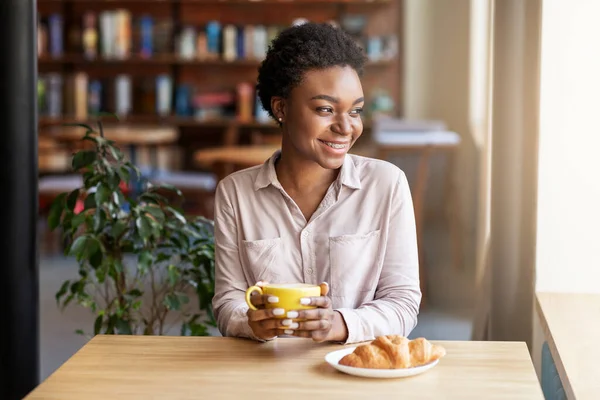 Chica negra alegre con taza de café fresco y delicioso croissant en la mesa en la cafetería — Foto de Stock