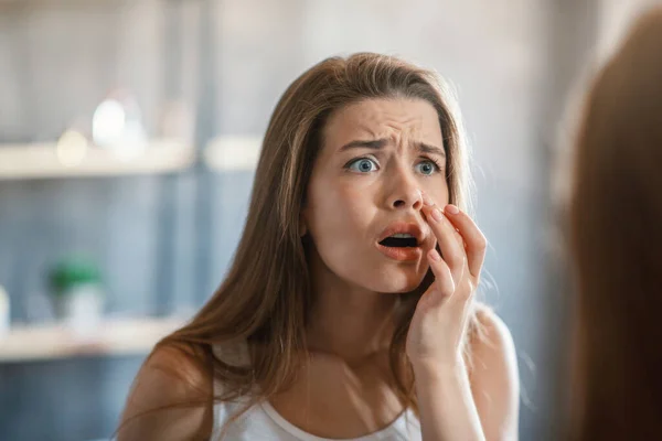 Mulher chocada tocando acne na bochecha enquanto olha no espelho em casa — Fotografia de Stock