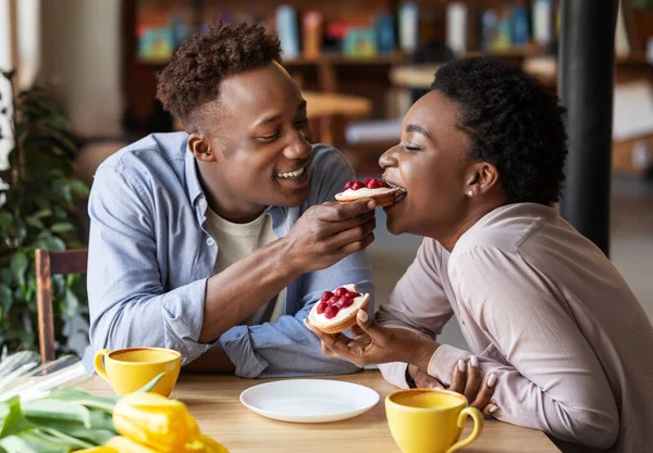 Millennial μαύρο άνδρα σίτιση νόστιμο τάρτα μούρο στην κοπέλα του σε ζεστό καφέ — Φωτογραφία Αρχείου