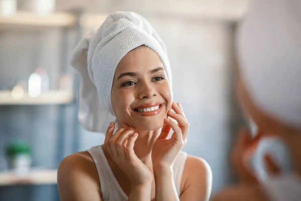 Attraente giovane donna con telo da bagno sulla testa che tocca la sua pelle luminosa vicino allo specchio a casa — Foto Stock