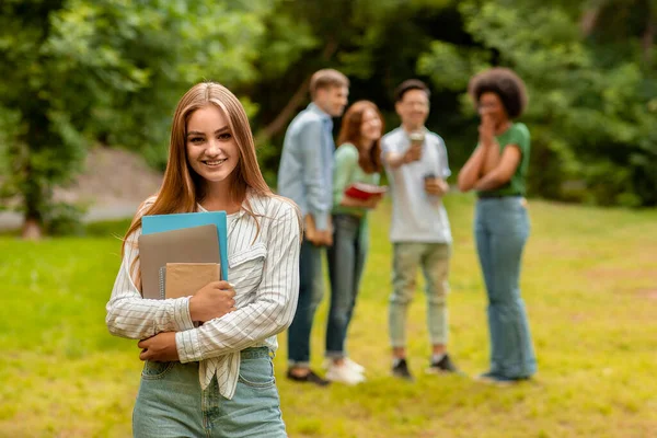 Lächelndes Millennial Student Girl hält Arbeitsbücher in der Hand und posiert im Freien mit Freunden im Hintergrund — Stockfoto