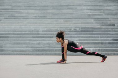 Açık havada antrenman yaparken esneme hareketleri. Kablosuz kulaklıklı Afrikalı Amerikalı kız merdivenlerde geriniyor.