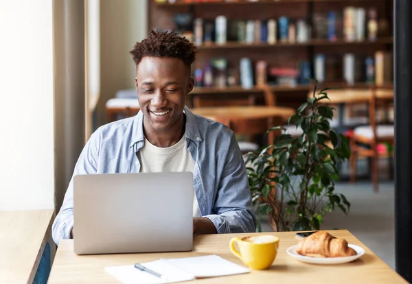 Chico negro feliz que trabaja en línea en el ordenador portátil en la acogedora cafetería de la ciudad, espacio libre — Foto de Stock