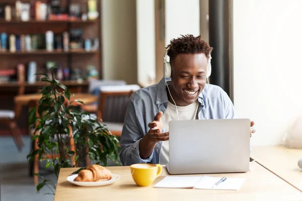コーヒーショップのラップトップコンピュータからオンラインで通信しているヘッドフォンの幸せな黒人男性、コピースペース — ストック写真