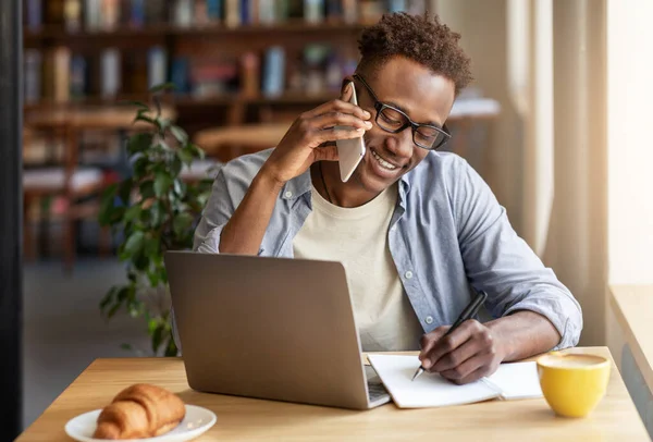 Chico negro alegre hablando en el teléfono inteligente mientras estudia en línea en la cafetería — Foto de Stock