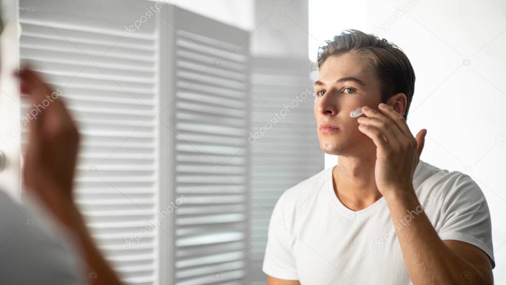 Handsome young man looking in the mirror applying cream