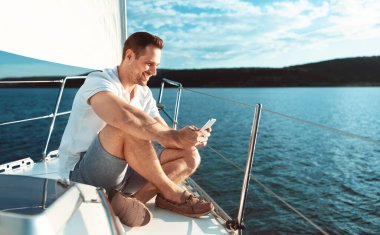 Akıllı telefon kullanan adam güvertede oturuyor açık denizde yelken açıyor.