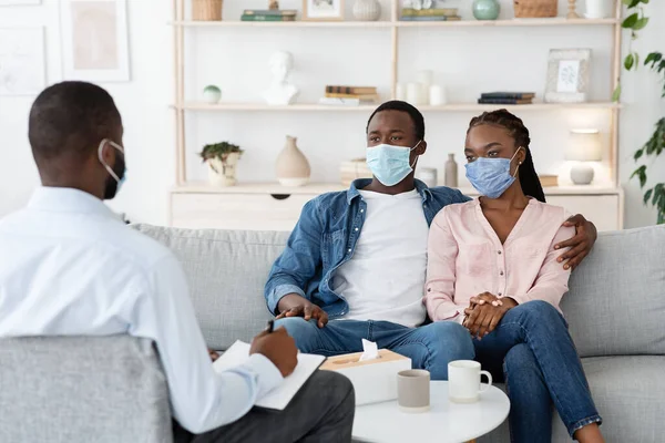 Terapeuta familiar hablando con una pareja negra sentada con máscaras protectoras en el sofá en la oficina —  Fotos de Stock