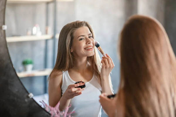 Ομορφιά και μακιγιάζ. Όμορφη νεαρή γυναίκα εφαρμογή blusher στο πρόσωπό της κοντά στον καθρέφτη στο σπίτι — Φωτογραφία Αρχείου