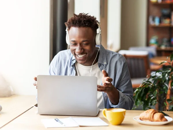 Online kommunikáció. Vidám afro-amerikai férfi headset beszél barátaival laptopon a városi kávézóban — Stock Fotó