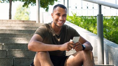 Parkta antrenman yaparken merdivenlerde cep telefonu kullanan neşeli siyah bir sporcu.