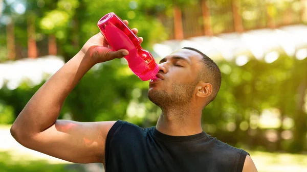 Hombre negro sediento bebiendo agua de la botella deportiva después de su entrenamiento en el parque — Foto de Stock