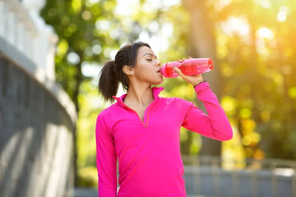Chica negra en sudadera rosa bebiendo agua durante el jogging — Foto de Stock