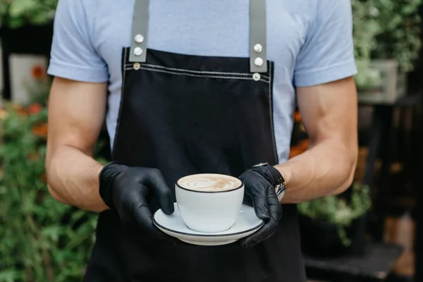 Garçom em avental e luvas, segurando cappuccino no café ao ar livre — Fotografia de Stock