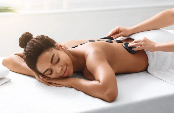 Therapist placing black hot stones on african lady back — Stock Photo, Image