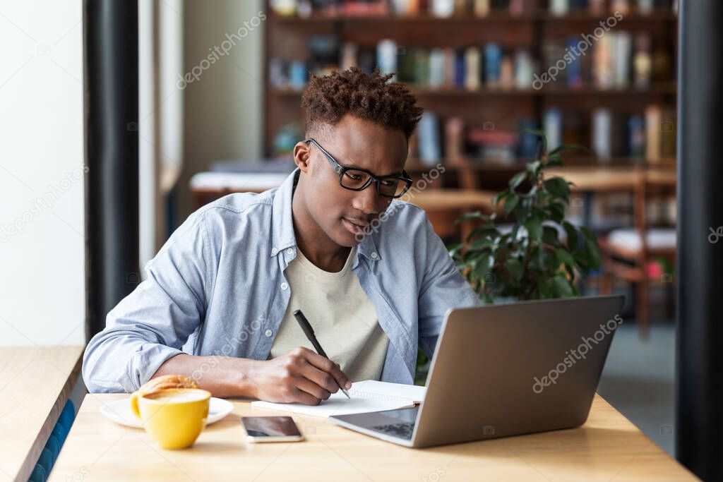 Serious black man taking notes while studying online in front of laptop at city cafe