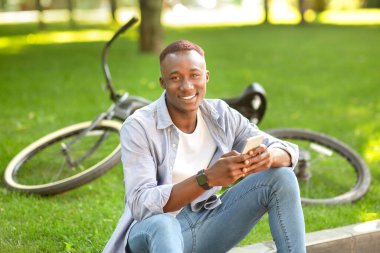 Parkta bisikletinin yanında oturan cep telefonlu gülümseyen siyah adam.