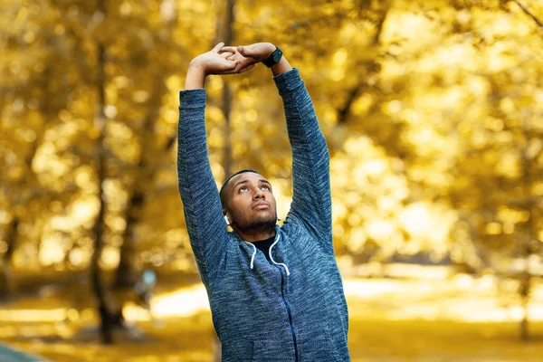 Milenial negro deportista estirando los brazos antes de correr en el parque en la mañana de otoño —  Fotos de Stock