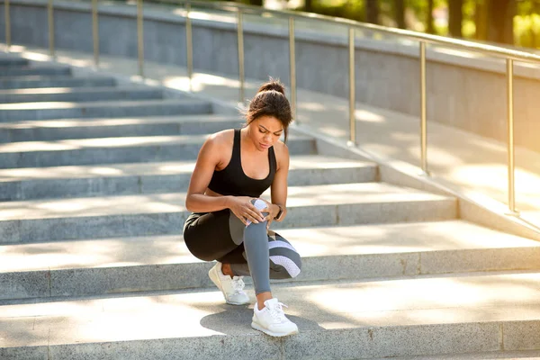 Chica negra en ropa deportiva sentado en las escaleras, masaje de rodilla — Foto de Stock