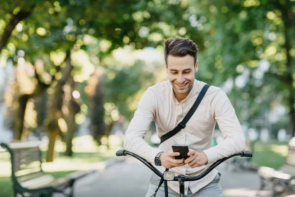 Redes sociais ao ar livre. Sorrindo cara atraente senta-se na bicicleta no parque e olha para o smartphone — Fotografia de Stock