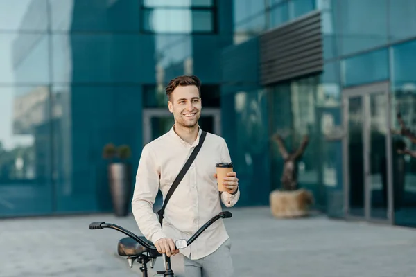 Café takeaway e eco transporte. Sorrindo homem segurando vidro e ir com bicicleta perto de prédio de escritórios — Fotografia de Stock