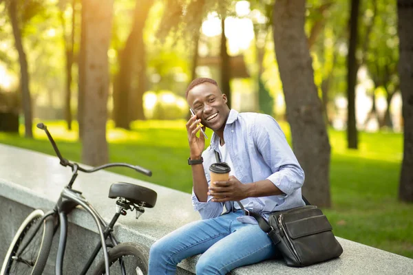 Homem negro alegre com café takeout falando no celular perto de sua bicicleta no parque — Fotografia de Stock