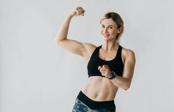 Motivación para hacer ejercicio. Mujer feliz en ropa deportiva con reloj inteligente demuestra bíceps y cuerpo muscular —  Fotos de Stock