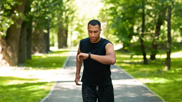 Atractivo chico negro mirando su rastreador de fitness mientras trota en el parque —  Fotos de Stock