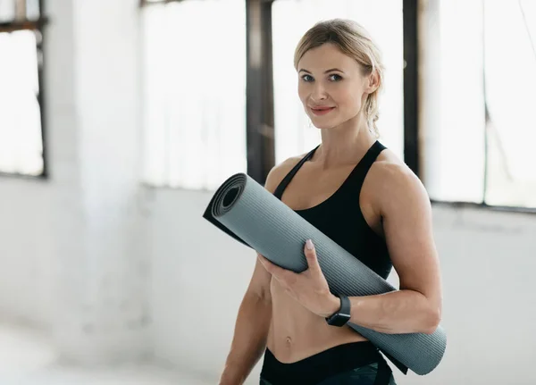 Retrato de treinador feminino muscular no ginásio em sportswear com tapete em suas mãos olha para a câmera — Fotografia de Stock