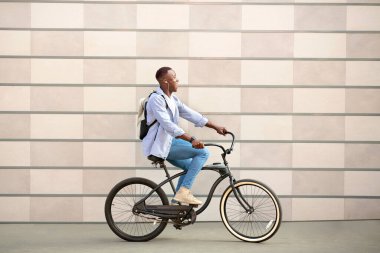 Sırt çantalı ve kulaklıklı heyecanlı siyah adam dışarıda tuğla duvarın yanında bisiklet sürüyor.