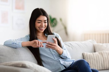 Sosyal ağlar. Gülümseyen Asyalı kız evdeki akıllı telefondan internete bakıyor.