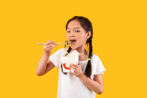 Chino chica comiendo fideos con palillos que sostiene la caja, fondo amarillo — Foto de Stock